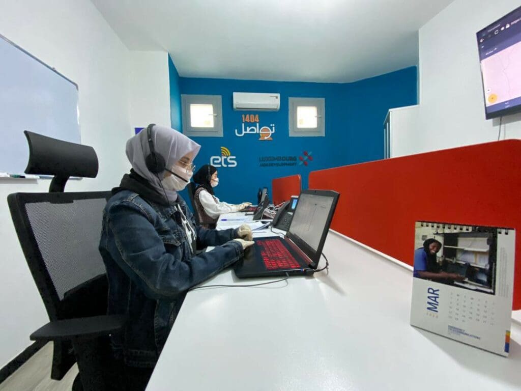 Two women in hijab and medical masks work at laptops in a call center
