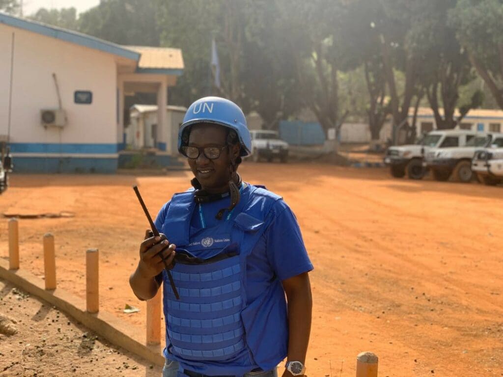 A man in blue UN protective gear and a helmet speaks into a walkie-talkie
