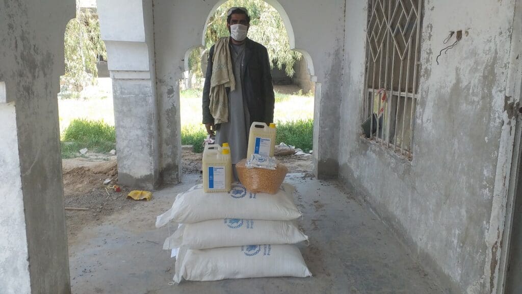 Obaidullah, a farmer wearing a mask, stands in front of a pile of food rations.