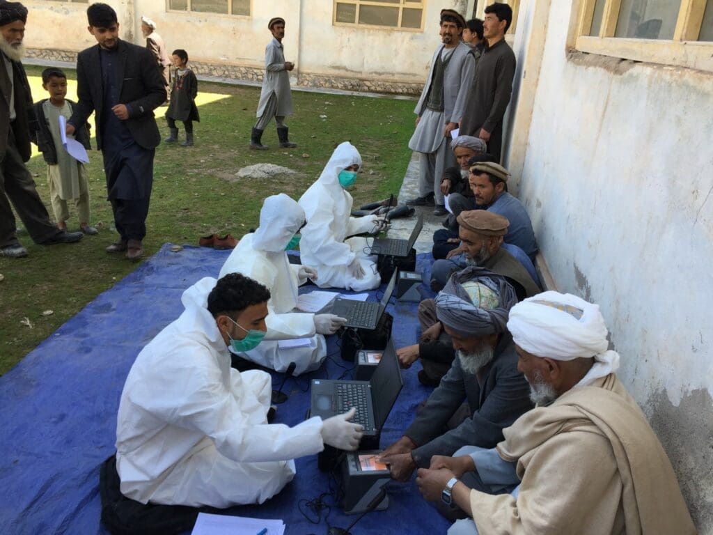 WFP workers in protective gear sit on a blue tarp, with laptops, across from Afghan people waiting for food assistance