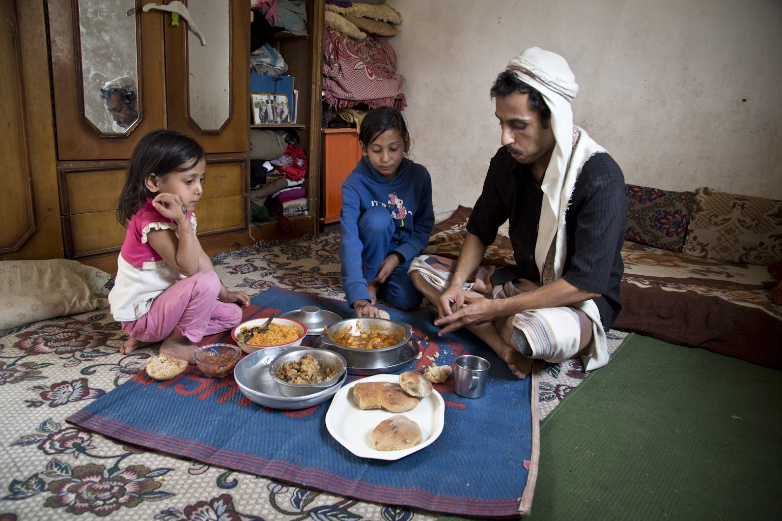 Walid and his two daughters sit on the floor with a blue mat covered in food dishes