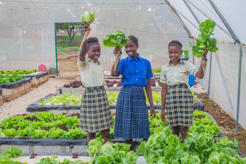 Three young women hold up veggies, standing in a garden.