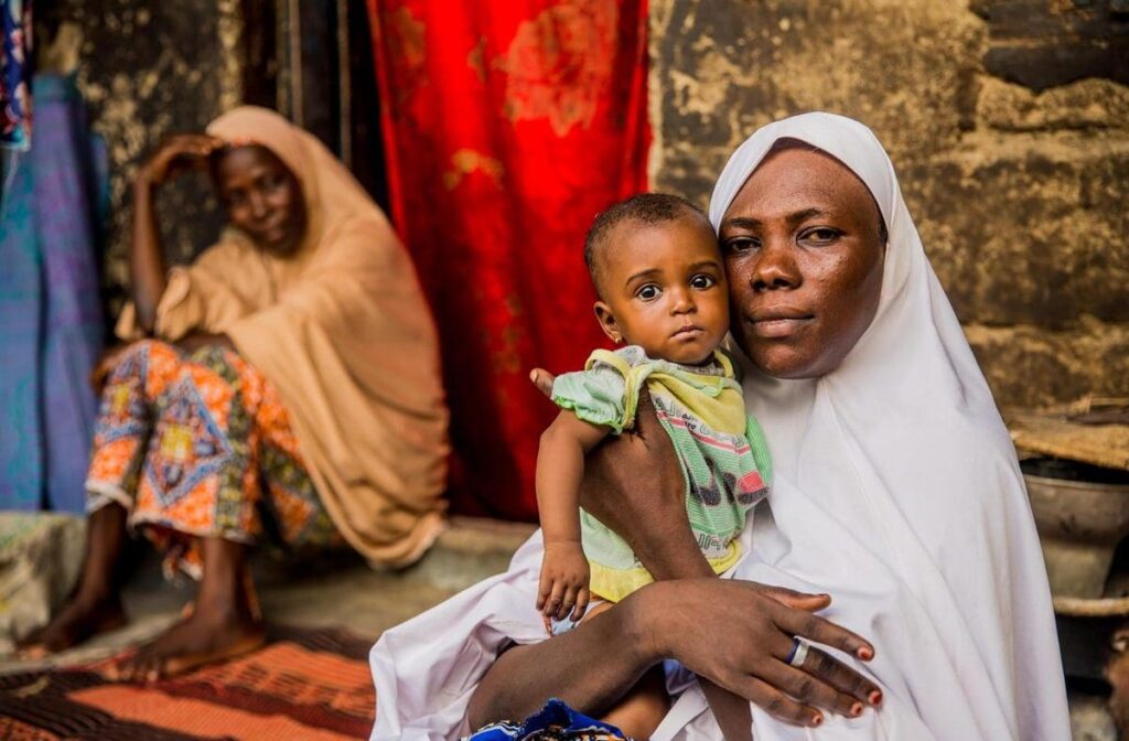 A mother sits, holding her child, with another woman nearby.