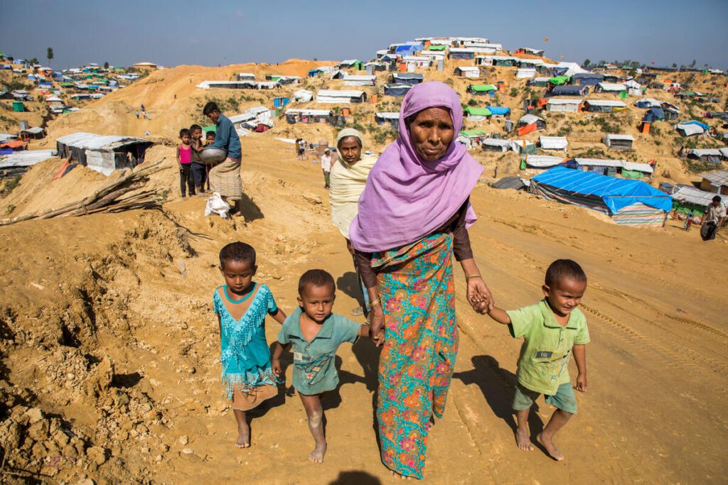 A grandmother in a headwrap walks with three of her grandchildren through a refugee camp.