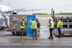 A group of WFP workers stands near a pallet of food near a cargo jet