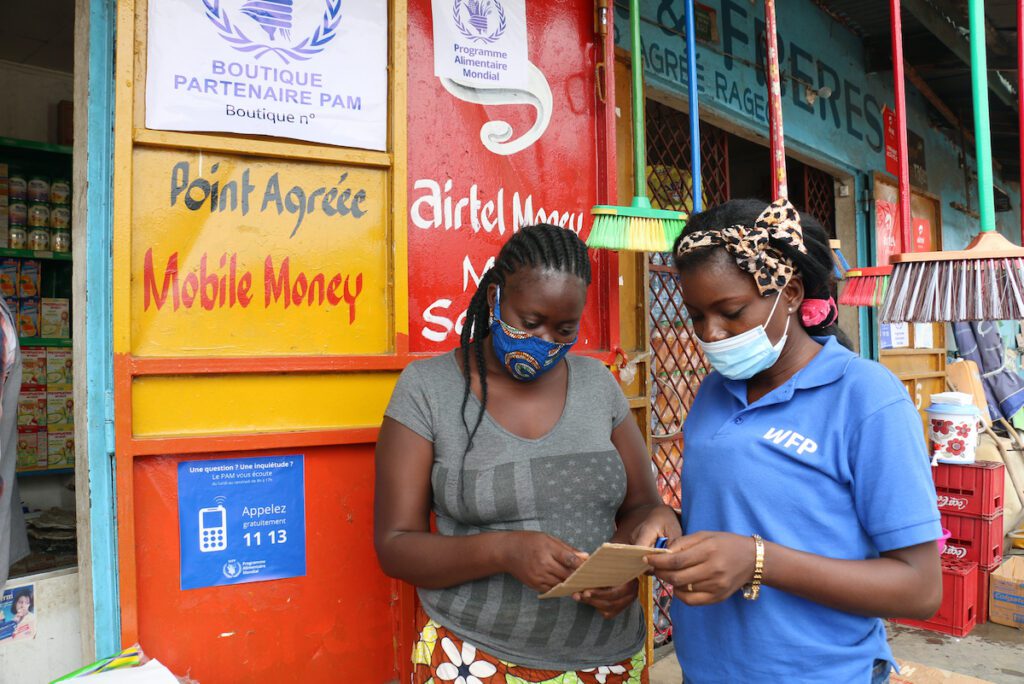 Two women in masks stand in front of a colorful kiosk store with a sign that reads "Mobile Money"