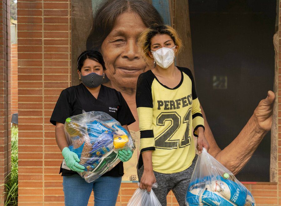 Two women wearing medical masks and holding bags of food are standing and looking at camera.