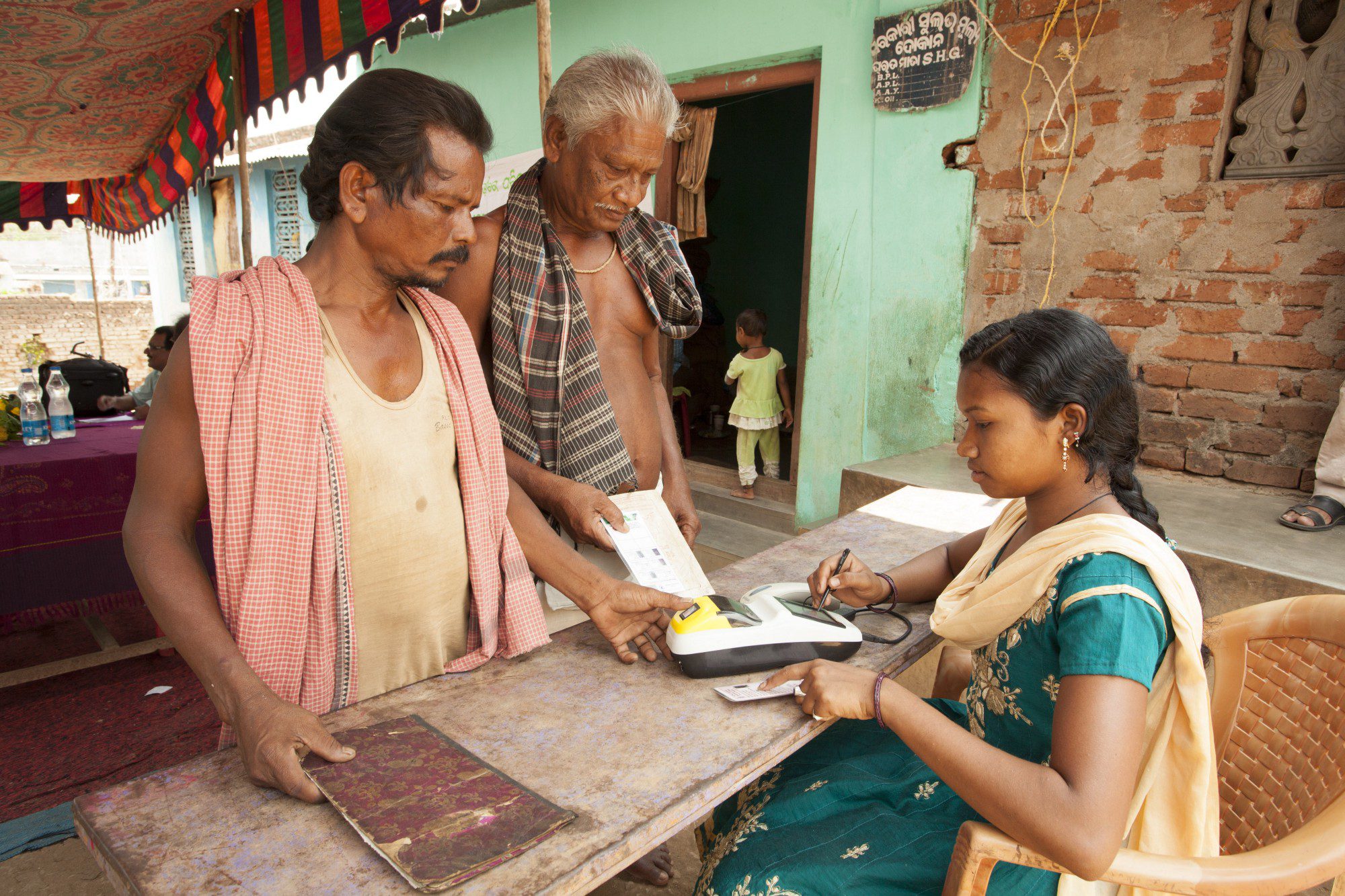 A woman in a sari sits at a table, two men stand in front of her.
