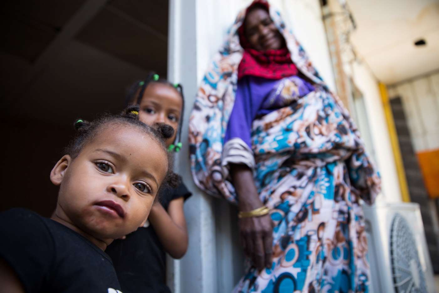 Two young girls stand in front of their home with their grandmother.