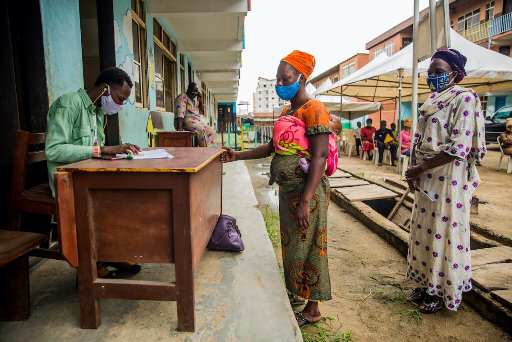 A woman in a mask with a baby wrapped to her back stands in front of a man sitting at a desk.