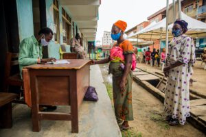 A woman in a mask with a baby wrapped to her back stands in front of a man sitting at a desk.