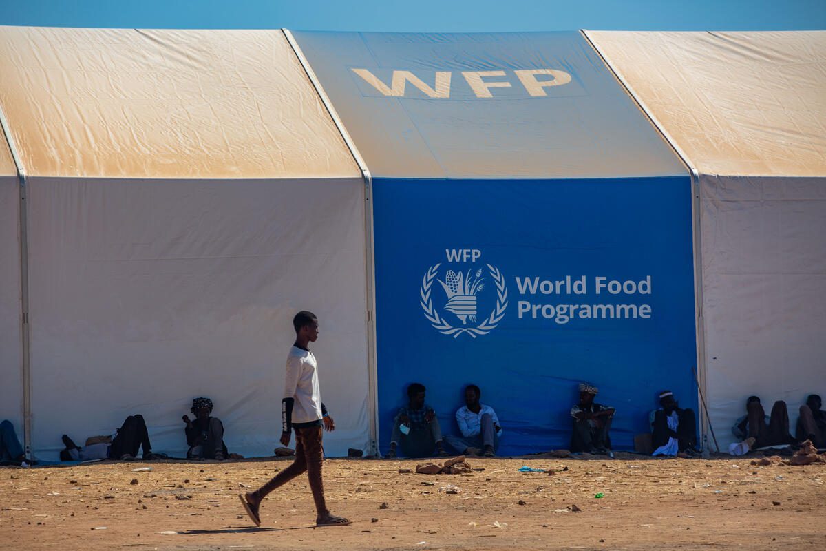 Refugees sit outside a WFP mobile storage unit. One man walks in front of them.