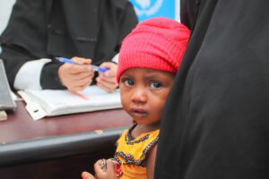 A young child sits on her mother's lap, looking back at the camera in a pink hat.