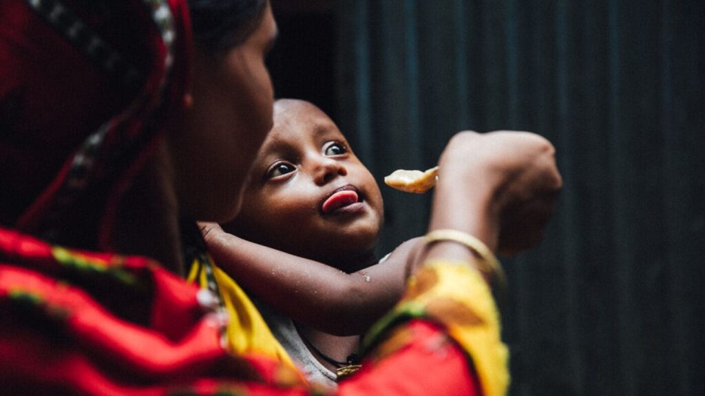 A mother feeds her child fortified porridge on a spoon, the child sticks out his tongue, ready to take a bite