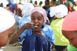 A young girl sits on the ground and eats a spoonful of food.