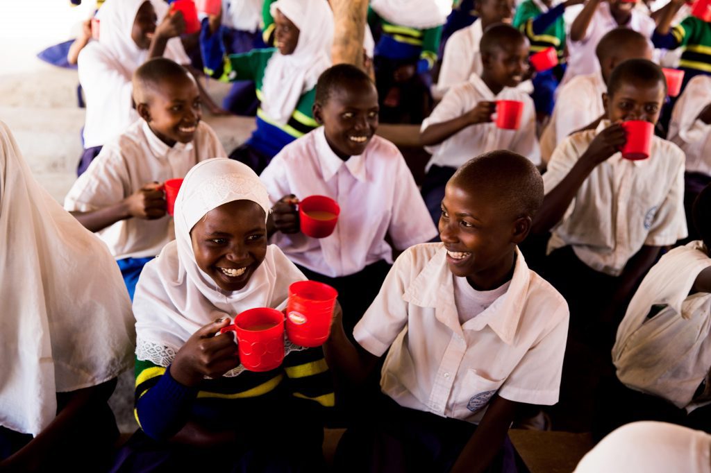 A girl smiles while eating her school meal, with her classmates surrounding her
