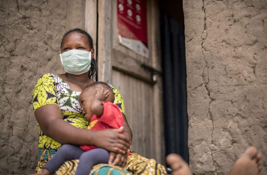 A mother wearing a mask holds her baby in her lap while sitting on the floor.