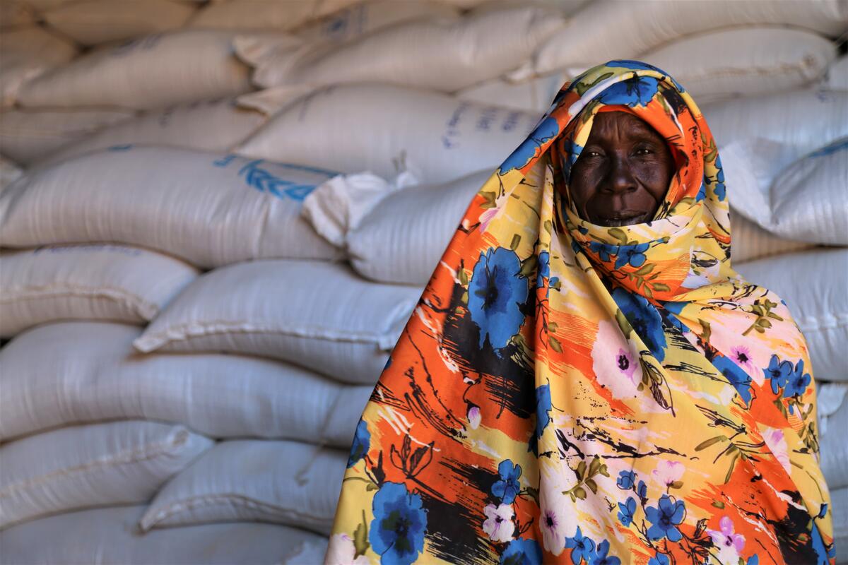 A woman in a colorful headwrap stands in front of a pile of WFP food bags.