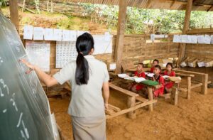 A teacher points at a chalkboard, standing in front of four young students.