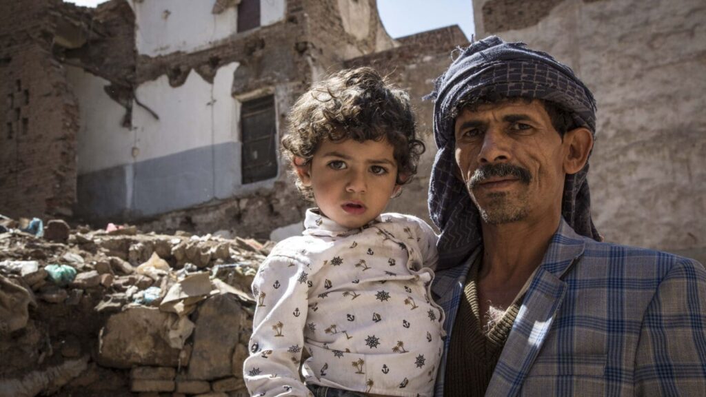 A man in a head scarf holds his daughter, standing in front of a burned out building.