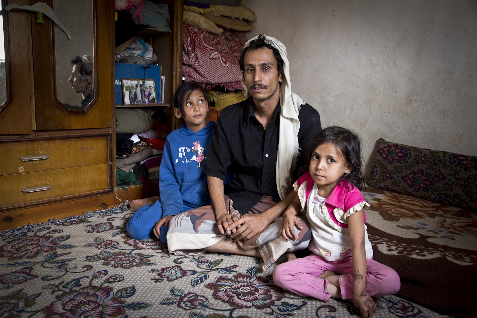 Walid sits with his two daughters on the floor, one daughter looks up at him