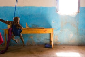 A baby hangs in a weighing sleeve in front of a blue wall.