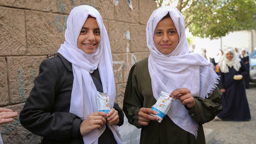 Two school girls in headwraps smile and eat snacks.