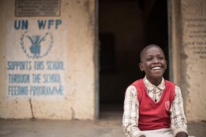 A young boy sits outside his school in a red vest
